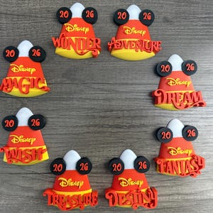 May include: Eight candy corn-shaped ornaments featuring black Mickey Mouse ears with the number 20 on the left and 26 on the right. Each ornament displays the word Disney in yellow and a unique name in red: Wonder, Adventure, Magic, Dream, Wish, Fantasy, Treasure, and Destiny.