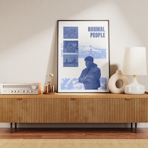 Puede incluir: Un póster enmarcado con el texto "NORMAL PEOPLE" e imágenes azules se exhibe en un mueble de madera. El mueble también contiene un equipo de música, una lámpara y jarrones decorativos. La habitación tiene una paleta de colores neutros.