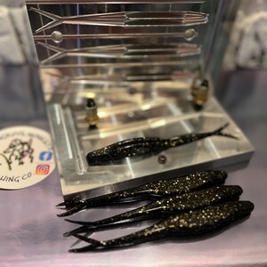 May include: A close-up shot of a fishing lure mold with several black and gold speckled lures. The mold is made of metal and has several cavities. A logo for "Mangrove Mafia Fishing Co." is in the background.