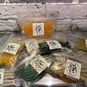 May include: A selection of soft plastic fishing lures in clear plastic bags. The lures are in various colours, including yellow, green, black, and brown. Each bag has a label with the text "Mangrove Monkeys Fishing Co." and a graphic.