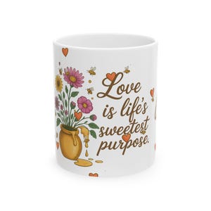 May include: White ceramic mug with a floral design, including pink, yellow, and purple flowers, a honey pot, and the text "Love is life's sweetest purpose." The design also features hearts and bees.