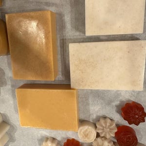 Puede incluir: Una variedad de jabones hechos a mano en varias formas y colores. Las barras rectangulares varían de beige claro a marrón dorado. Los jabones más pequeños tienen forma de flor y son redondos, en tonos blanco, beige y naranja rojizo. Los jabones están dispuestos sobre una superficie clara.