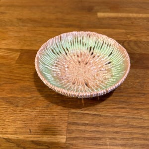 May include: A small, round decorative bowl with a textured surface. The bowl features a color gradient, transitioning from a light green rim to a peach center. The bowl is placed on a wooden surface.