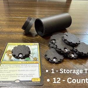 May include: Black storage tube with a screw-on lid, alongside a Magic: The Gathering card and several black counters. The counters have white numbers. The text on the card reads "Kumena, Tyrant of Orazca".