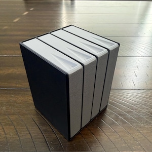 May include: A black storage case containing four vertical slots with light gray textured dividers. Each slot features a white top edge with a recessed triangular indentation. The unit is rectangular and holds the four dividers upright.