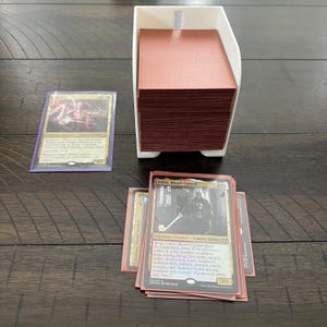 May include: A white card holder with a stack of red-edged cards, and several individual cards with illustrations and text. One card is labeled "Odric, Blood-Cursed". The cards are on a dark wooden surface.