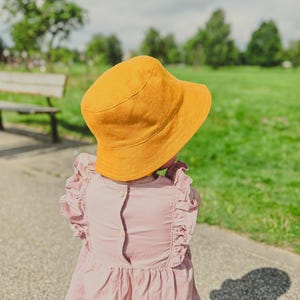 Op de afbeelding: Een mosterdgele bucket hat gedragen met een oudroze mouwloze top met ruches bij de schouders en twee kleine knoopjes op de rug. De hoed heeft een brede, afhangende rand en een ronde kroon, terwijl de top een geplooide taille heeft.