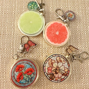 Peut inclure: Quatre porte-clés ronds avec des cadres en métal doré. Les motifs comprennent une tranche de citron vert, une tranche de pamplemousse, des coquelicots rouges et un motif floral en vitrail avec une abeille. Chaque porte-clés a un mousqueton et un petit charme.