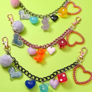 May include: Three charm bracelets with a variety of colourful charms. Each bracelet has a chain with a lobster clasp, a fluffy ball, a glitter heart, and small charms like hearts, bears, and dice. The background is a bright green.