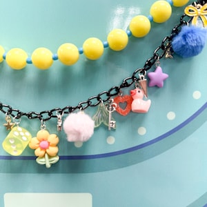May include: Two necklaces with colorful charms. The top necklace has yellow beads. The bottom necklace has a black chain with charms including a pink star, a duck, a heart, a key, a flower, a dice, and a pom-pom.