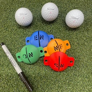 May include: Three white Titleist golf balls with the number 1 in red. Four colored golf ball alignment tools are labeled with the letters EN, NE, SN, and ON. A black and silver Sharpie marker is also in the image.