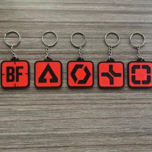 Könnte beinhalten: Fünf Schlüsselanhänger mit roten quadratischen Anhängern und silbernen Schlüsselringen. Jeder Anhänger hat ein anderes schwarzes grafisches Design. Ein Anhänger hat die Buchstaben "BF". Die Schlüsselanhänger werden auf einer Holzoberfläche präsentiert.