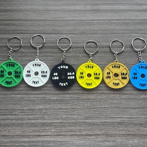 Op de afbeelding: Zes kleurrijke gewichtplaat sleutelhangers in groen, wit, zwart, geel en blauw. Elke sleutelhanger heeft een zilveren ring en toont de tekst "YOUR", "45 LBS", "20.4 KGS" en "TEXT".