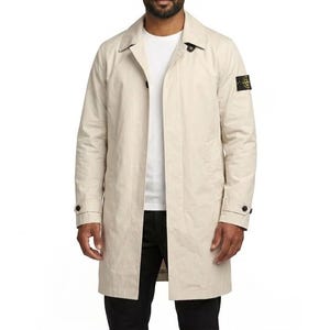 May include: A beige trench coat with a button-down front and a small square patch on the upper arm. The coat is worn over a white t-shirt and black pants. The coat has a classic, tailored design.