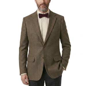 May include: A brown tweed blazer with a subtle plaid pattern. The blazer features a notched lapel, two front pockets, and a burgundy bow tie. The person is wearing a light-colored button-down shirt and dark gray pants.