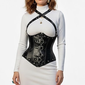 May include: Black leather corset with silver-tone buckles and studded details. The corset features a structured design with a halter-style harness. The garment is worn over a white long-sleeved dress, creating a contrast in textures and colors.