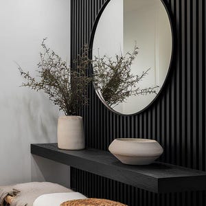 May include: Interior design featuring a black slatted wall with a round mirror. A black shelf holds a vase with dried branches and a decorative bowl. A woven bench with a cushion and a round woven pillow completes the scene.