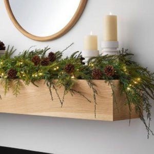 May include: A wooden mantelpiece adorned with a green garland, pine cones, and fairy lights. Two cream-coloured candles stand on white candle holders. A round mirror hangs above the mantelpiece, reflecting the scene.