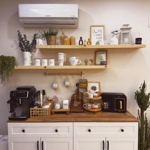 May include: A well-organized coffee station with a white cabinet, wooden countertop, and floating shelves. The shelves display mugs, decorative items, and a framed sign that reads "Coffee is always a good idea." Appliances include a coffee maker and a toaster.