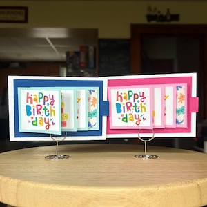 May include: Two accordion-style greeting cards featuring the text "happy birthday" in colorful, playful lettering. One card is mounted on a dark blue base, and the other on a bright pink base, both supported by silver wire stands on a wooden surface.
