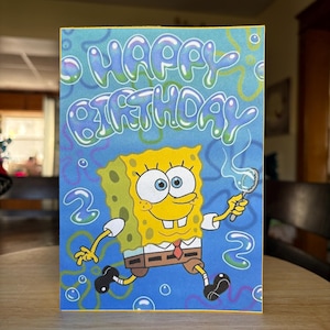 May include: A colorful birthday card featuring SpongeBob SquarePants blowing bubbles. The card has a blue background with the words "HAPPY BIRTHDAY" in bubble letters. SpongeBob is yellow with brown shorts.