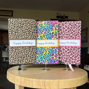 May include: Three rectangular greeting cards featuring animal print patterns. The first has a brown and black leopard print, the second a multicolored neon leopard print, and the third a pink and black leopard print. Each card displays the text Happy Birthday.