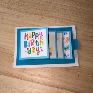 May include: A handmade birthday card with a teal border and a white base. The front features the words "Happy Birthday" in colorful letters. Inside are several small, decorated panels with illustrations.