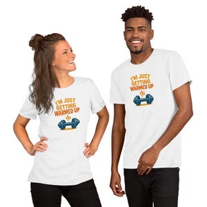 May include: Two people wearing white t-shirts. Each shirt has the text "I'M JUST GETTING WARMED UP" in orange and blue, along with a blue dumbbell graphic with steam. The shirts are a fitness-themed design.