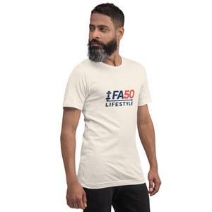 May include: A cream-coloured t-shirt featuring the text "FA50 LIFESTYLE" in navy blue and red. The shirt has a crew neck and short sleeves. The model is wearing black trousers.