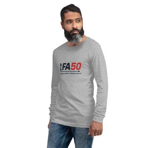 May include: A light grey long-sleeved shirt featuring the text "iFA50" in black and red. The phrase "Tactical Fitness. Powerful Results." is printed below. The shirt is paired with blue jeans.