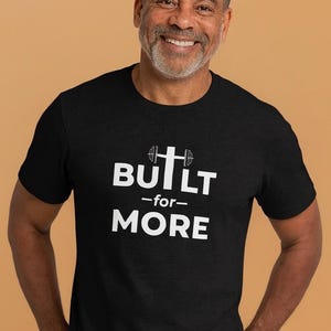 May include: Black t-shirt with the white text "BUILT -for- MORE". The word "BUILT" has a barbell graphic in place of the letter "I". The person in the image is smiling.