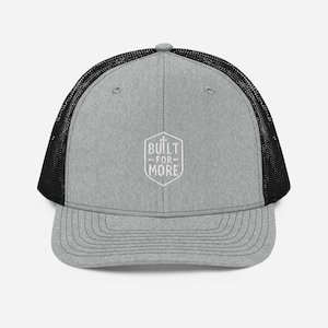 May include: Grey trucker cap with a black mesh back. The cap displays the text "BUILT FOR MORE" in a white, geometric design. It has a curved peak and a snap closure for adjustment.