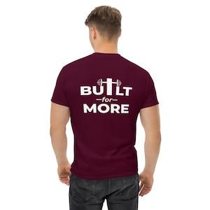 May include: A maroon t-shirt with the text "BUILT -for- MORE" in white. The design includes a barbell graphic above the word "BUILT". The shirt is made of a soft material and is suitable for casual wear.