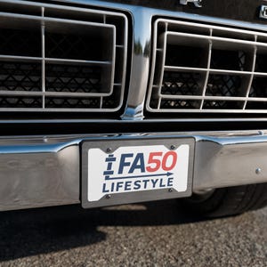 May include: A vehicle license plate displaying "FA50 LIFESTYLE" in blue and red, set within a silver frame. The plate is mounted on a chrome bumper, with the car's front grill and asphalt visible in the background.