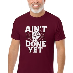 May include: A maroon t-shirt with the text "AIN'T DONE YET" in white, featuring a fist graphic. The shirt is short-sleeved and has a crew neck.