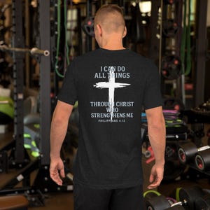 May include: Dark grey short-sleeved t-shirt featuring the text "I CAN DO ALL THINGS THROUGH CHRIST WHO STRENGTHENS ME" and a white cross graphic. The text is in a white, stylized font. The shirt is made of a soft material.