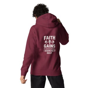 May include: A maroon hoodie featuring a white graphic on the back with the words "FAITH GAINS", a barbell and cross symbol. The text below reads "MY FAITH IS STRONG... I EXERCISE IT DAILY". The hoodie has a drawstring hood and a front pocket.