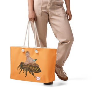 May include: Orange tote bag with a whimsical illustration of a baby riding a giant bee. The bag has white rope handles and a small square logo in the lower right corner. The bag is held by a person wearing tan pants and brown shoes.