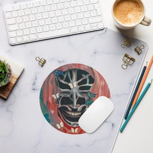 May include: Round mousepad featuring a surreal design with a skeletal face, red and blue butterflies, and a red backdrop. A white computer mouse sits on the pad, with a keyboard and coffee cup in the background. The mousepad is on a white marble surface.