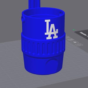 Könnte beinhalten: Ein leuchtend blauer Becherhalter mit dem weißen "LA"-Logo. Der zylindrische Becherhalter hat ein geriffeltes Band und einen Griff. Das Design ist schlicht und modern, ideal für Sportfans.