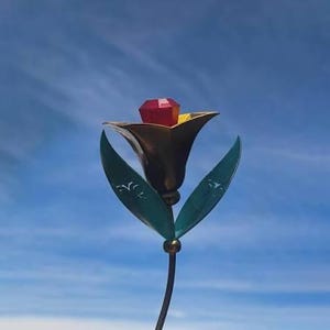 May include: A metal flower sculpture featuring a dark bronze-toned bloom with a faceted red gemstone at its center. The flower has two pointed teal-colored leaves with etched white patterns on a thin metal stem, set against a blue sky background.