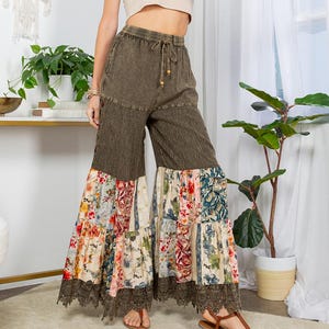 Puede incluir: Una mujer con un top corto beige, pantalones palazzo verde oliva con patchwork floral y un sombrero de paja. Los pantalones tienen cintura con cordón y ribete de encaje. El atuendo se combina con sandalias. Un atrapasueños cuelga en el fondo.
