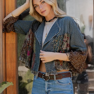Könnte beinhalten: Jeansjacke mit floralen und gemusterten Stoffeinsätzen. Die Jacke hat einen ausgefransten Saum und wird mit einer blauen Jeans und einem braunen Gürtel getragen. Die Ärmel sind ausgestellt und mit einem Blumenmuster versehen. Das Model trägt eine Halskette und Armbänder.