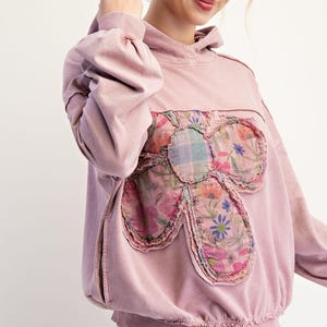 May include: A dusky pink hoodie featuring a large floral appliqué. The flower is crafted from assorted floral fabrics with a frayed edge. The hoodie includes a drawstring hood and side pockets. The sleeves have a gathered detail.