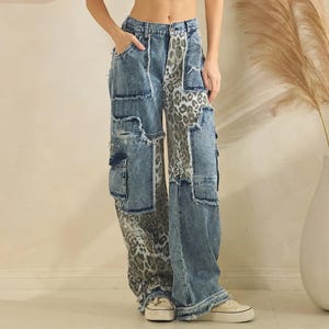 May include: Light wash denim cargo trousers with leopard print panels and distressed details. The trousers have multiple pockets and a wide leg fit. The model is wearing a white crop top and canvas trainers.