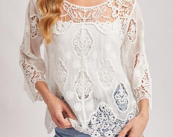 May include: White, long-sleeved blouse with intricate lace detailing on the neckline, sleeves, and bodice. The blouse has a relaxed fit and is paired with denim shorts. A straw hat completes the casual, bohemian look.