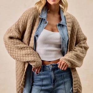 Könnte beinhalten: Eine Frau trägt eine beige Strickjacke über einer hellblauen Jeansjacke und einem weißen gerippten Crop-Top. Sie trägt auch blaue Jeans. Die Strickjacke hat ein strukturiertes Muster. Das Outfit ist lässig und stilvoll.