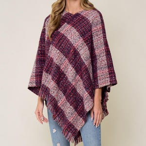 May include: A burgundy, pink, and gray striped poncho with fringe detail. The poncho is worn with a black hat and blue jeans. The V-neck poncho has a textured weave.