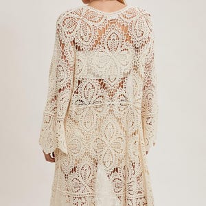 May include: Cream-colored, open-weave crochet cardigan with long sleeves and a scalloped hem. The cardigan features a floral pattern and is worn with a brown hat. This lightweight, bohemian-style garment is perfect for layering.