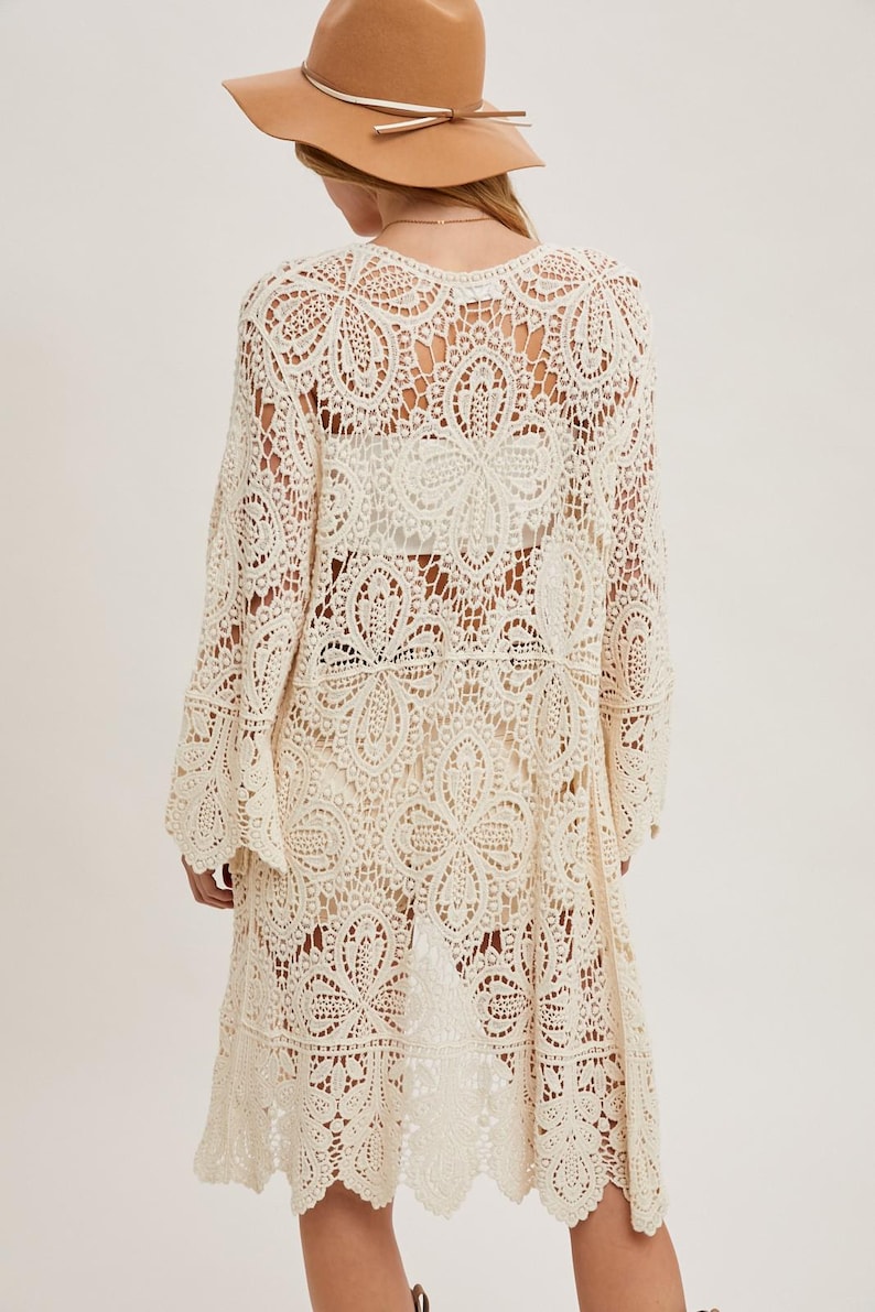 May include: Cream-colored, open-weave crochet cardigan with long sleeves and a scalloped hem. The cardigan features a floral pattern and is worn with a brown hat. This lightweight, bohemian-style garment is perfect for layering.